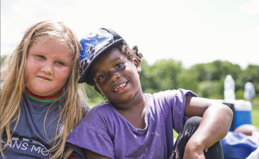 Photo of two girls playing outdoors. One is wearing a blue baseball cap and a purple t-shirt and smiling. The other is staring off-camera and is wearing a blue t-shirt with writing on it.