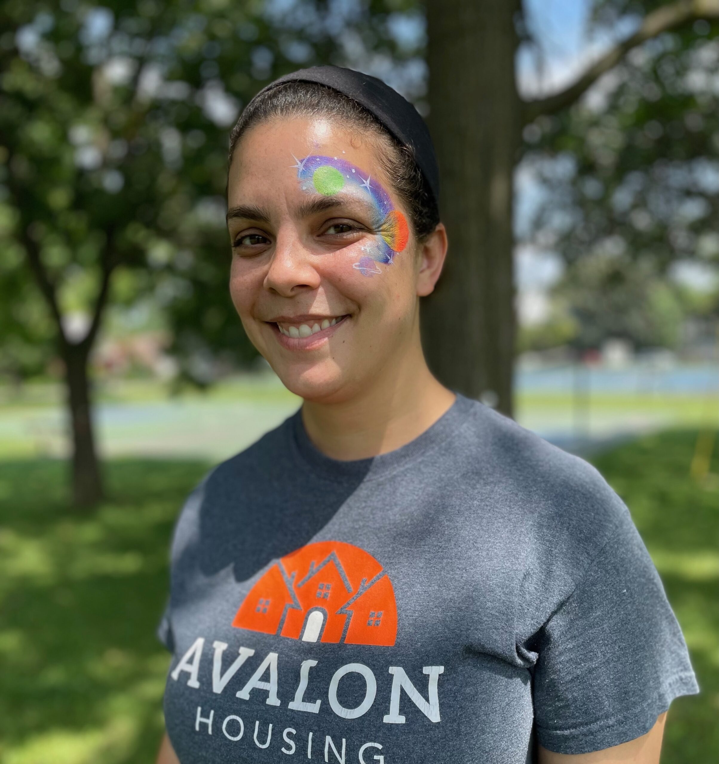Photo of a woman with black hair pulled up/back with a black hairband. She is smiling. Her face has a space galaxy with stars and planets painted on the side of her face around her left eye. She is wearing a charcoal grey Avalon t-shirt with an orange and white house logo and the name Avalon Housing on it in white.