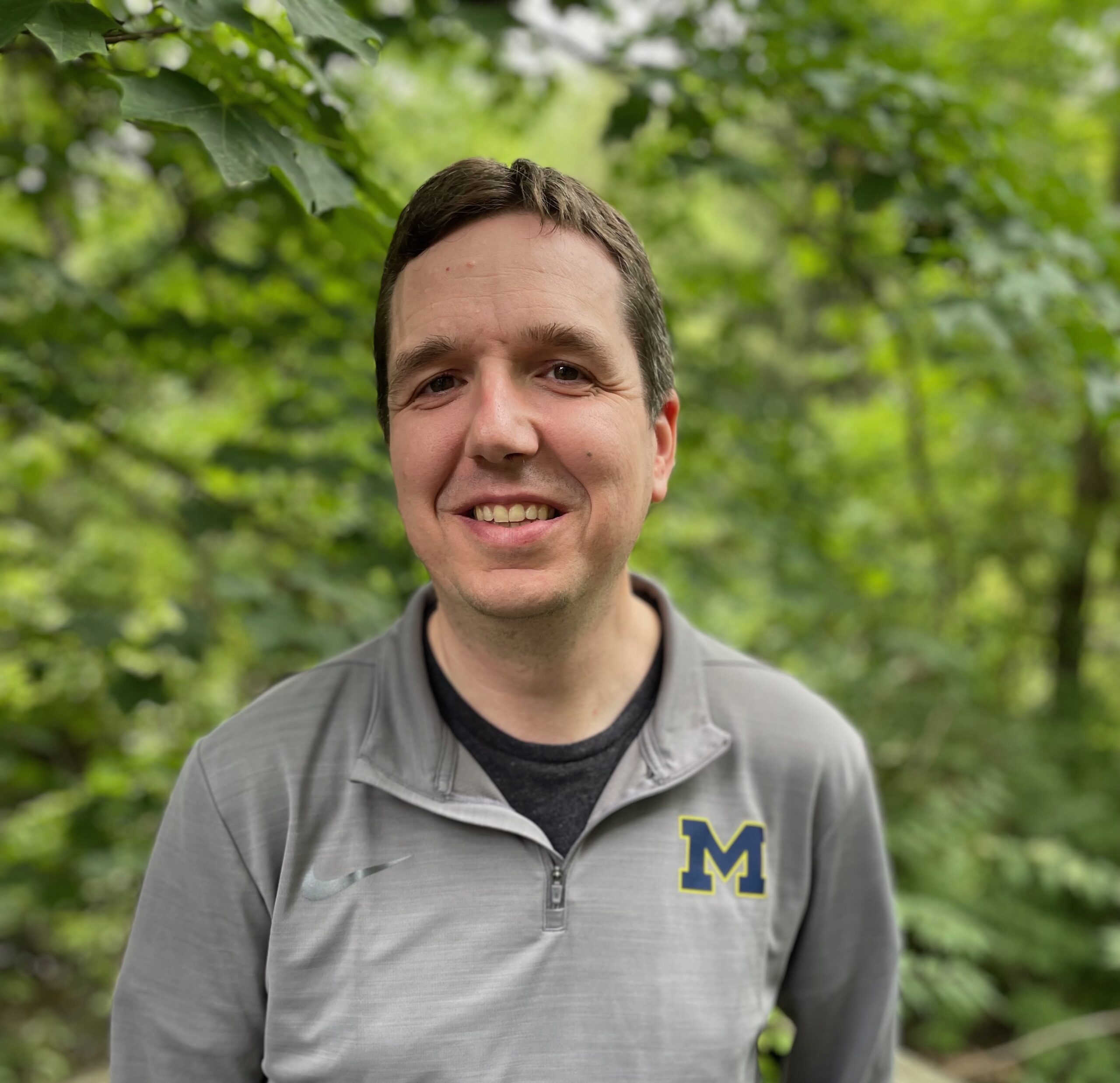 Photo of a man with short brown hair, smiling, wearing a grey zipped-front shirt with a University of Michigan navy blue block M trimmed in yellow, over a black t-shirt.