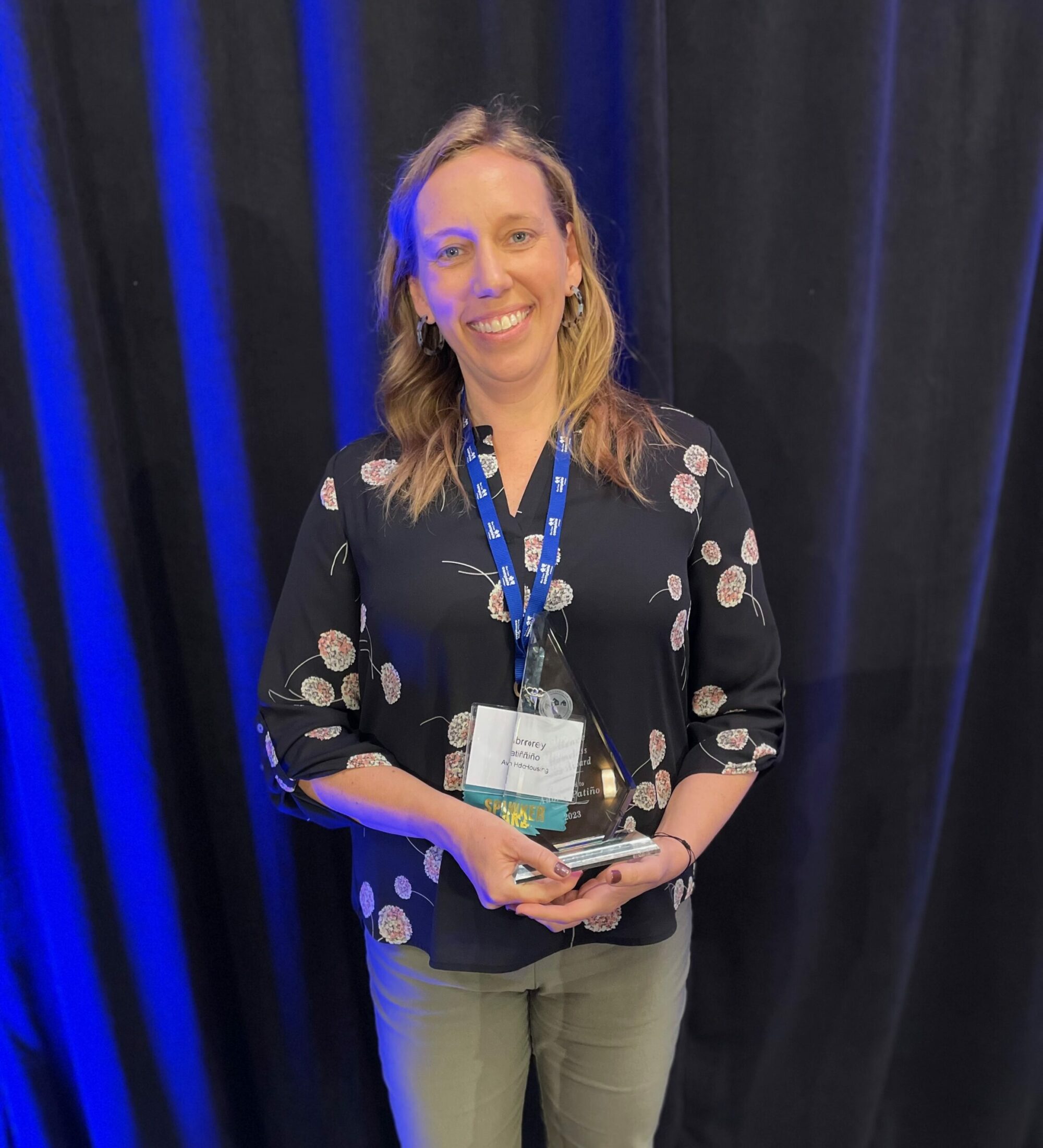 Photo of woman in a dark shirt with flowers, standing in front of a blue curtain, holding an acrylic award, entitled "2023 Excellence in Homeless Services Award"