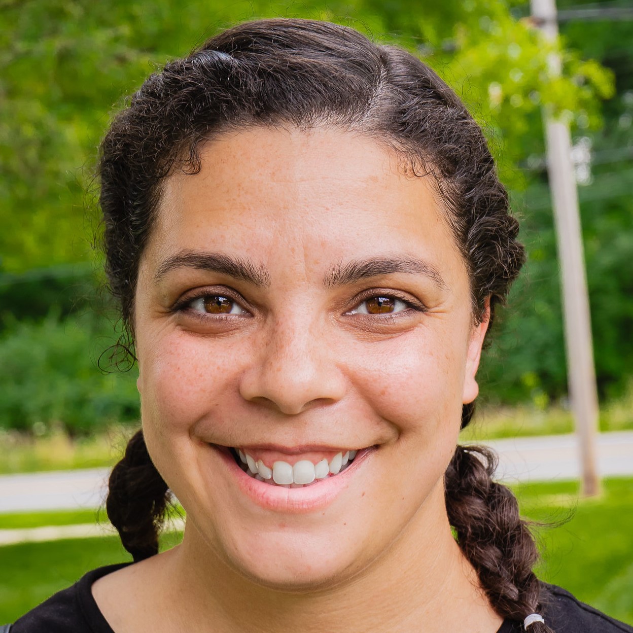 Photo of a woman with black hair pulled up/back with a black hairband. She is smiling. Her face has a space galaxy with stars and planets painted on the side of her face around her left eye. She is wearing a charcoal grey Avalon t-shirt with an orange and white house logo and the name Avalon Housing on it in white.